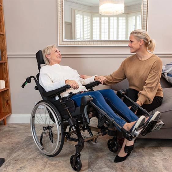 Altere Dame im Multifunktionsrollstuhl, junge Frau auf Couch daneben