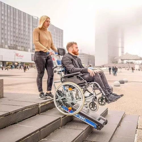 Frau hilft Mann im Rollstuhl mit Treppenraube eine Treppe auf einem öffentlichen Platz hinauf